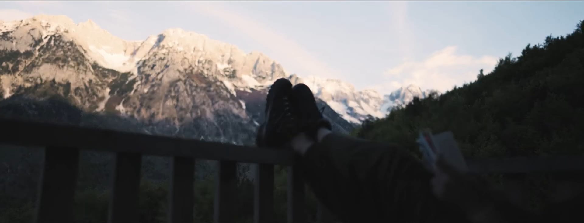 Still from a travel film in Albania: boots resting on a railing with a book, snow-capped mountains behind