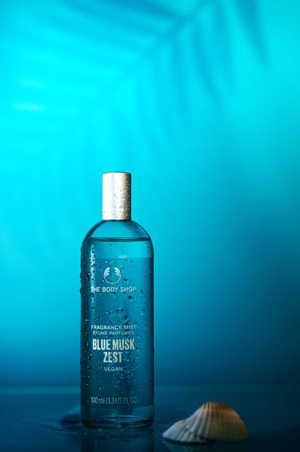 The Body Shop Blue Musk Zest bottle on a cyan set with a palm shadow and a seashell