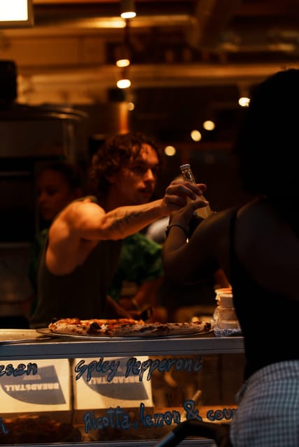 Pizza counter with staff passing a bottle under tungsten light